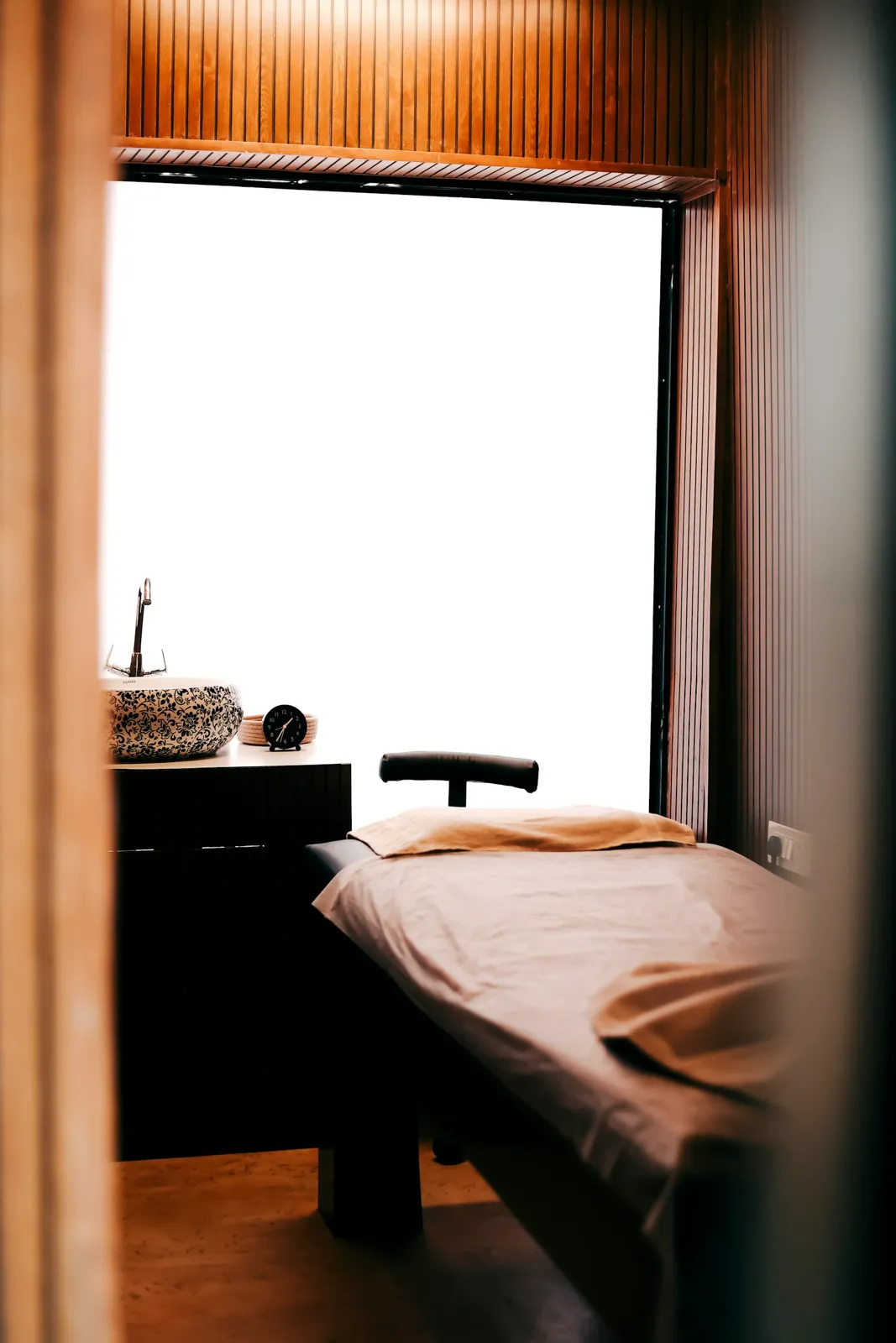 A calm treatment room with a massage bed and a basin, lit by a soft window.